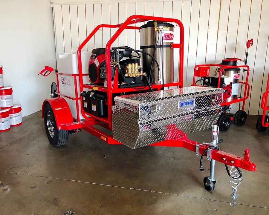 Hotsy hot-water machine in a red powder-coated frame on a single-axle trailer with a diamond-plate toolbox, beside buckets of Hotsy Breakthrough detergent